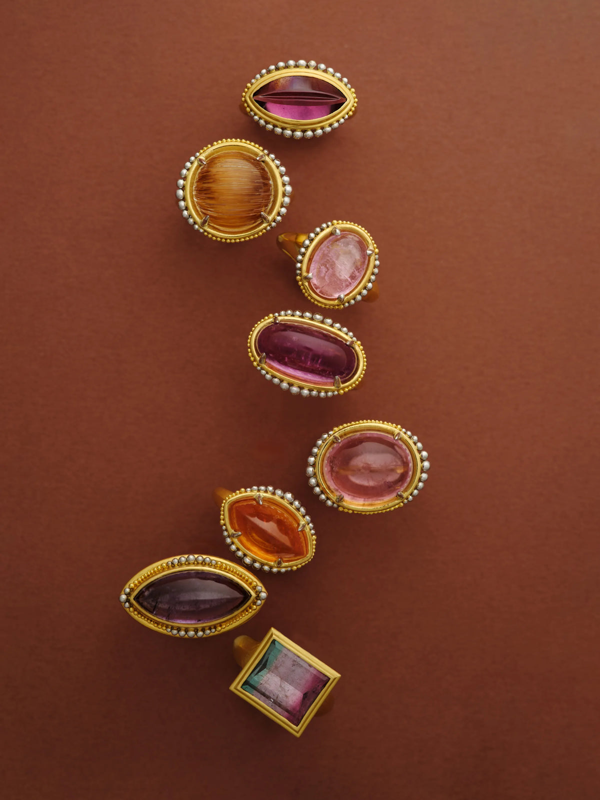 Seven 22k gold rings set with garnet, citrine, pink tourmaline, watermelon tourmaline, and amethyst cabochons, arranged in a row on a terracotta background