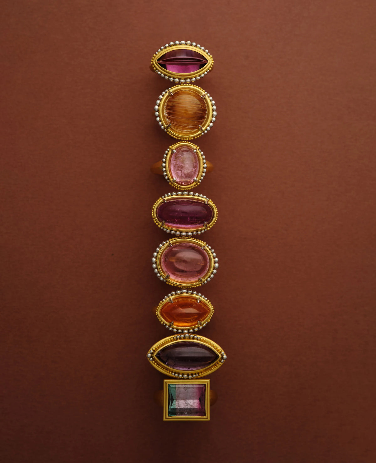 Eight 22k gold rings in a vertical row, set with garnet, citrine, pink tourmaline, watermelon tourmaline, and amethyst cabochons on a terracotta background
