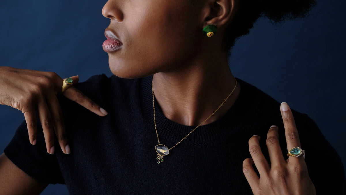 A woman wearing a gold emerald ring, a green tourmaline earring, and a gold intaglio pendant necklace with dangling gemstone drops