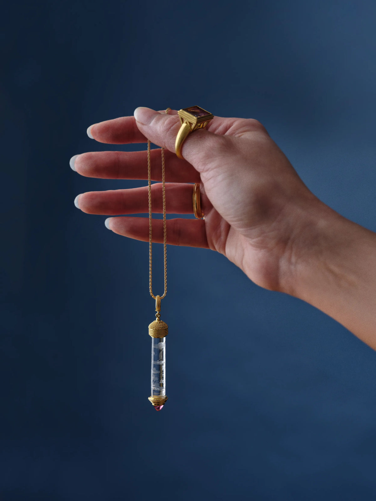 A hand wearing a gold ring set with a rectangular gemstone holds a gold chain necklace with a carved rock crystal vessel pendant tipped in pink tourmaline