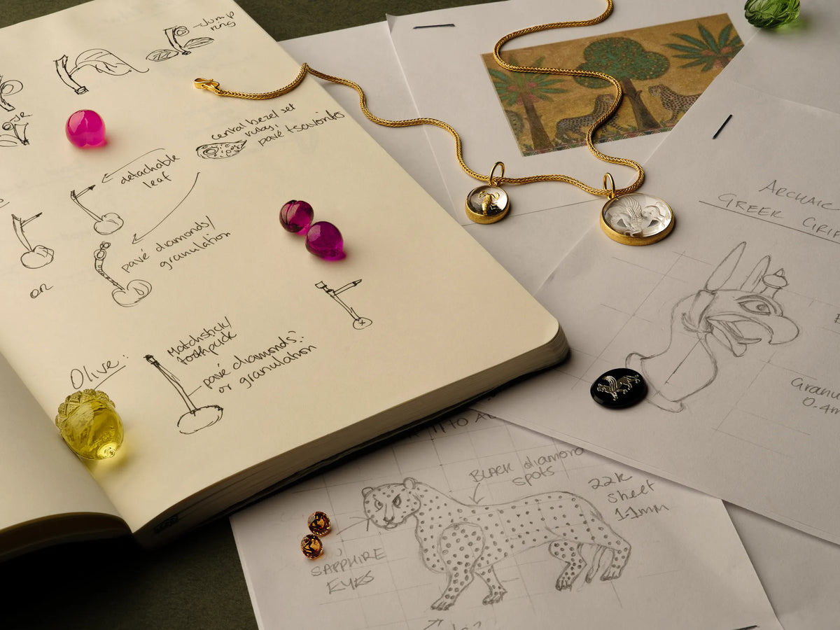 Open design sketchbook with hand-drawn jewelry notes, ruby cabochons, and gold pendants engraved with cheetah and griffin motifs inspired by antiquity