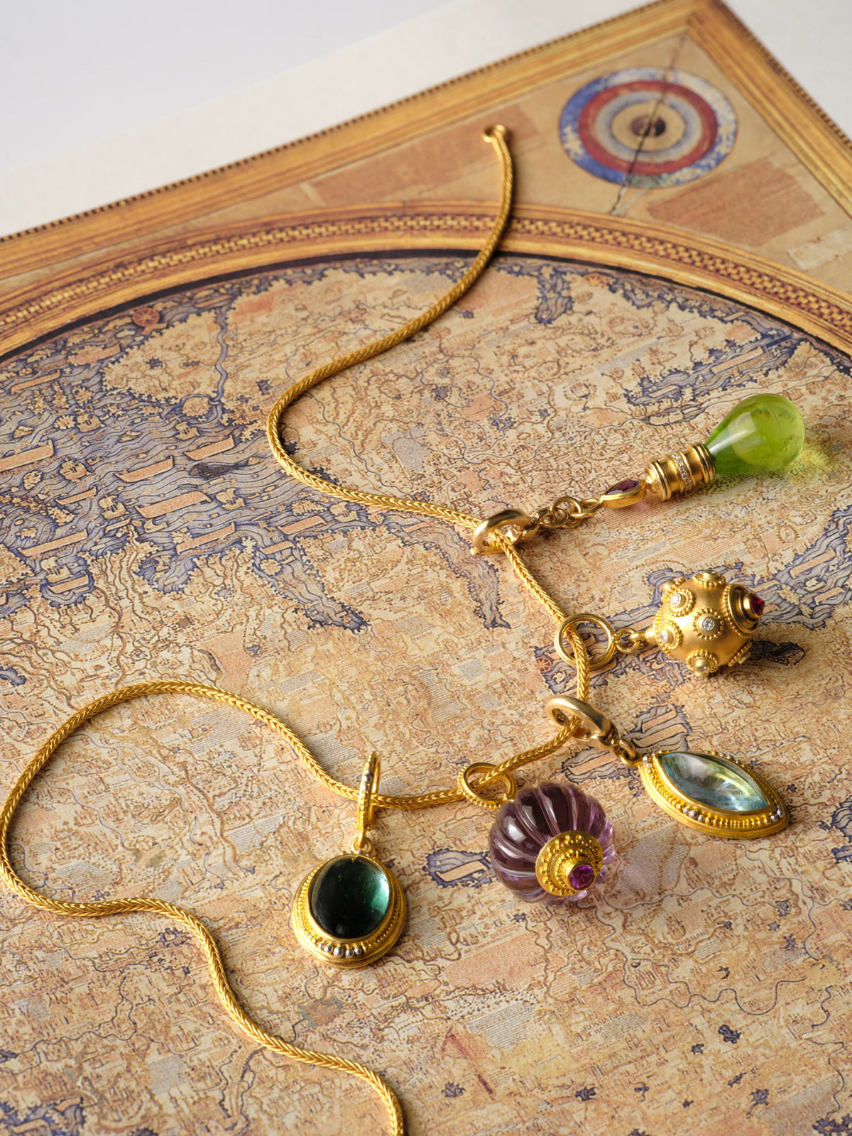 A 22k gold chain necklace with carved amethyst, peridot, tourmaline, aquamarine, and granulated gold charm drops, laid on an antique map