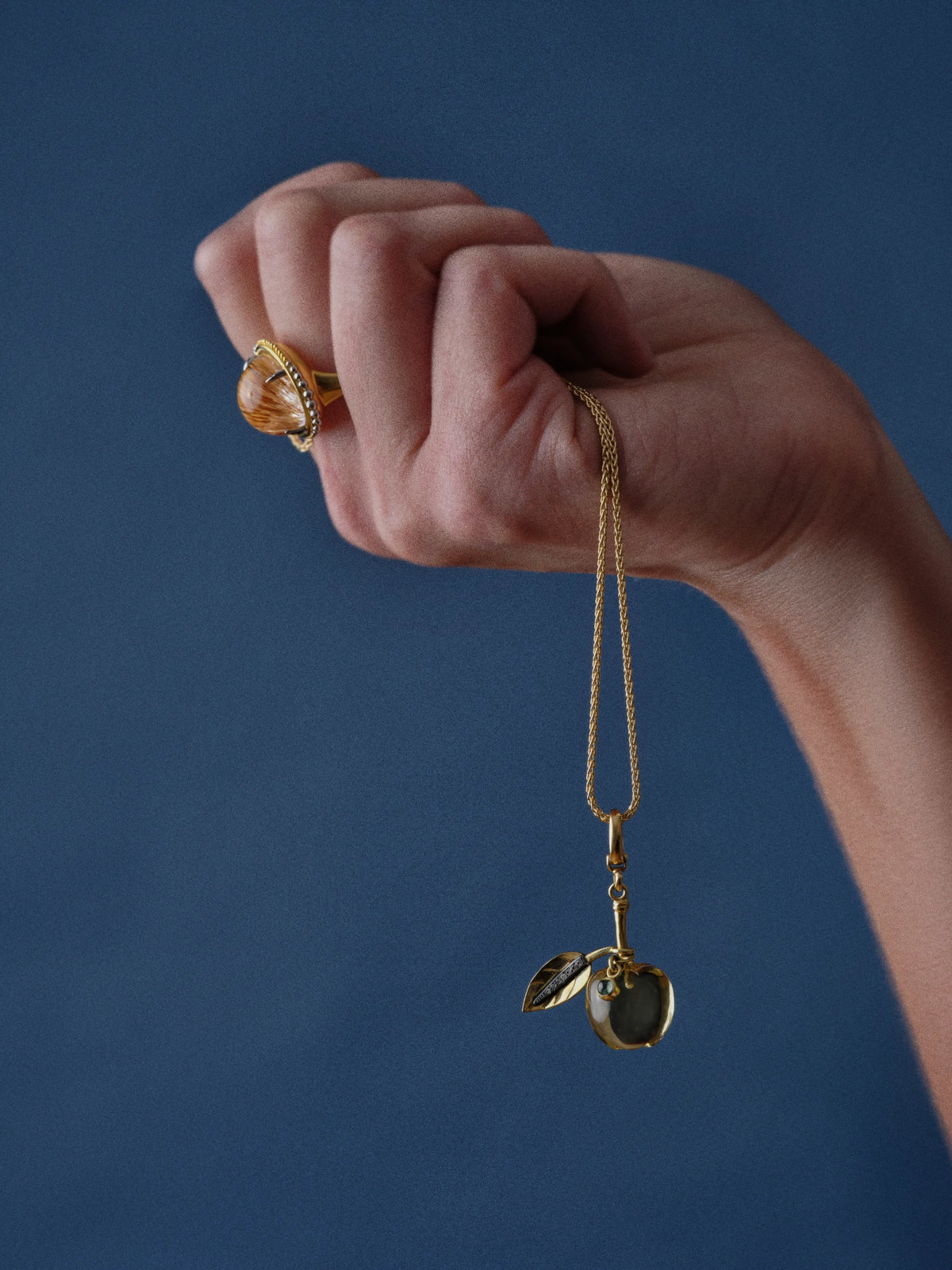 A hand wearing a citrine cabochon gold ring holds a gold chain necklace with a pavé diamond leaf and gemstone apple charm