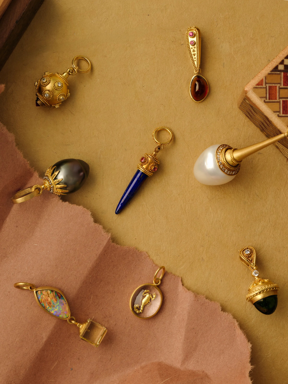 An assortment of 22k gold charm pendants set with garnet, lapis lazuli, opal, tourmaline, a Tahitian pearl, and a scorpion intaglio, arranged on tan and pink paper