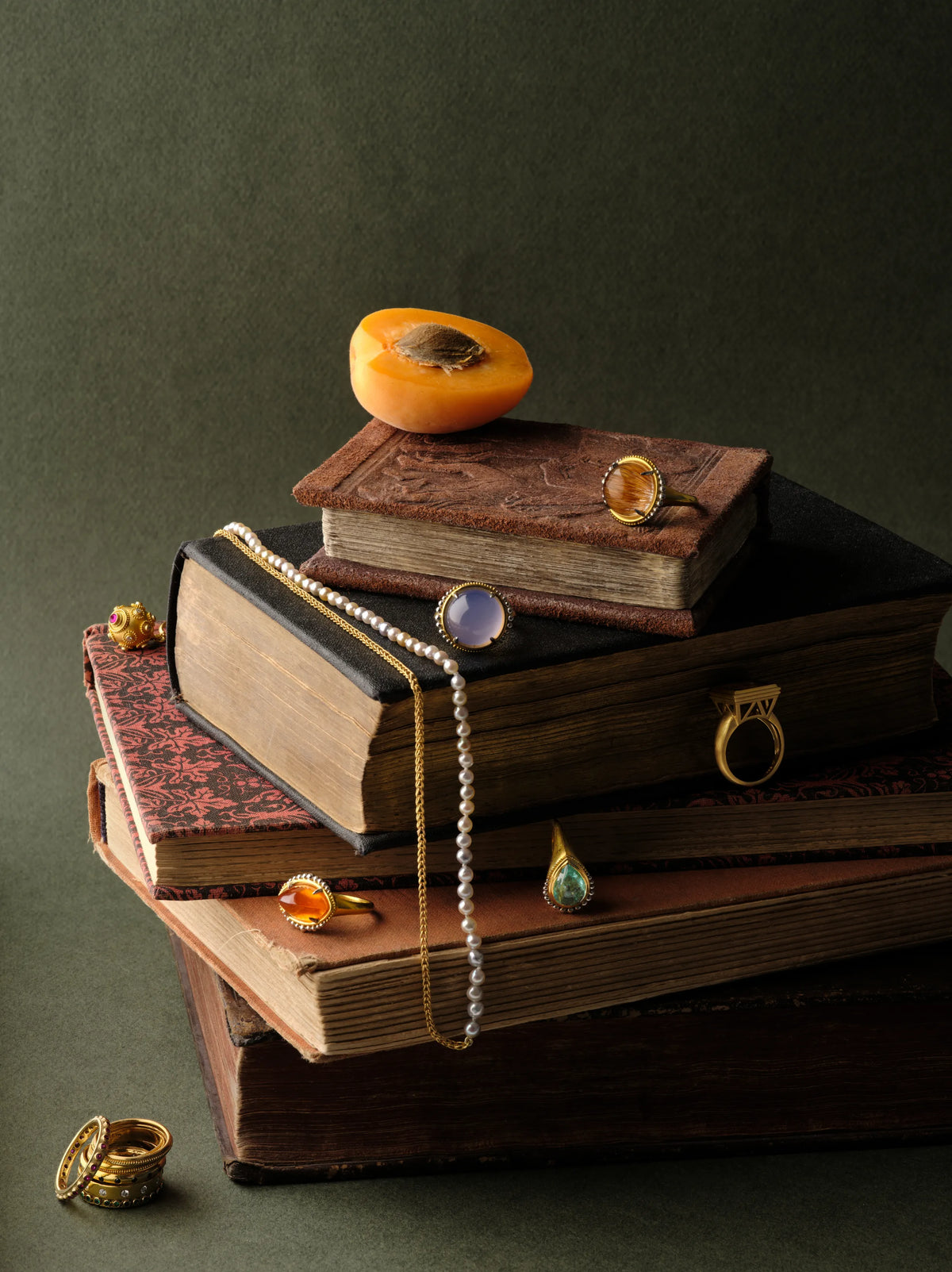 22k gold rings set with rutilated quartz, chalcedony, citrine, amber, and turquoise, styled on a stack of antique books with a pearl necklace and a halved apricot
