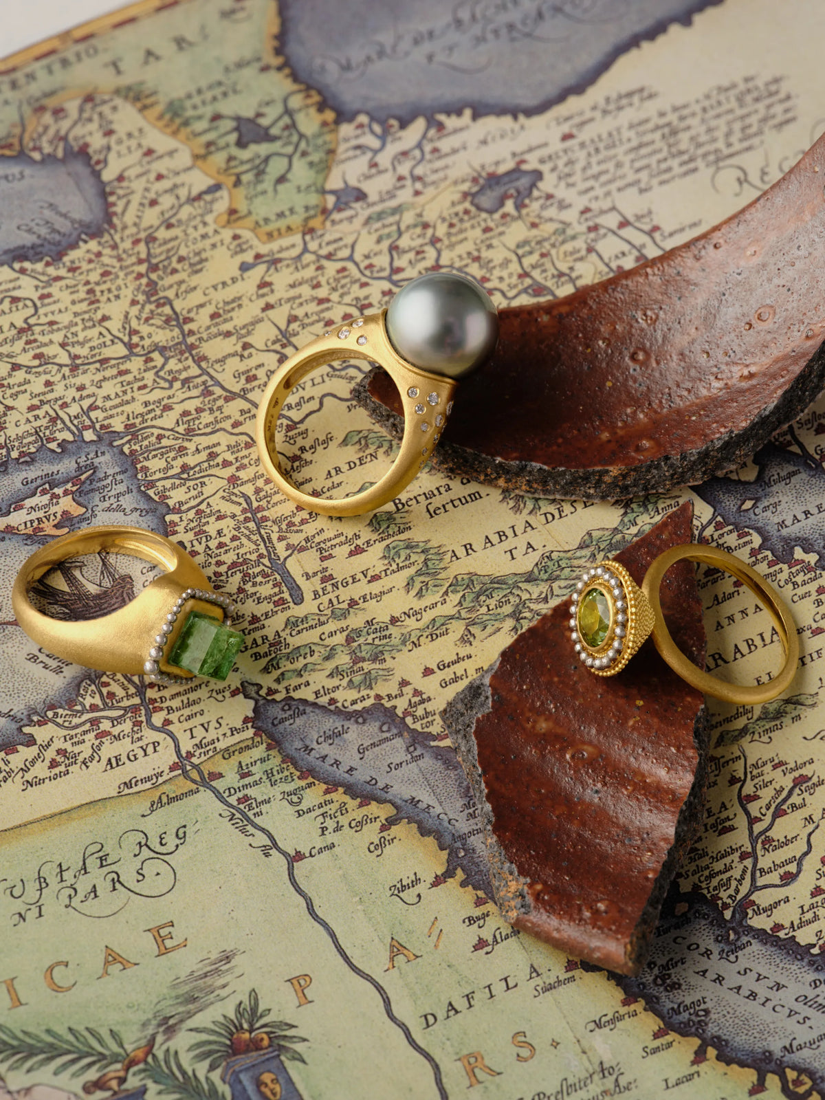 Four 18k matte gold rings with a Tahitian pearl, emerald, tourmaline, and diamond pavé, styled on an antique map with a terracotta shard.