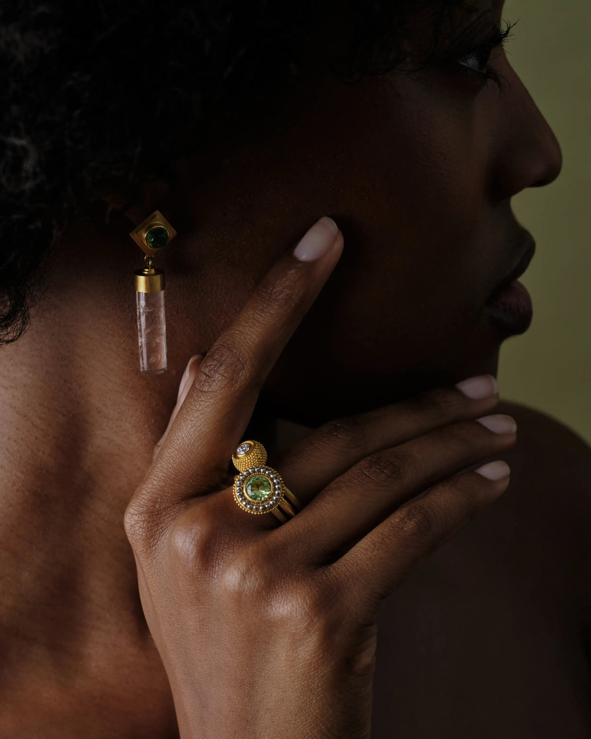 Close-up of a gold ring stack with peridot and diamond pavé, worn alongside a rock crystal drop earring against bare skin.
