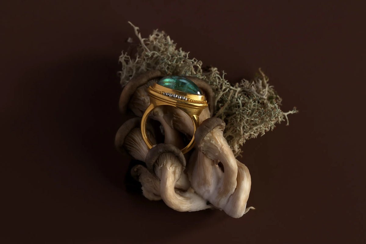 A 22k gold ring set with a cabochon tourmaline and pavé diamond band, styled among wild mushrooms and lichen against a dark brown background