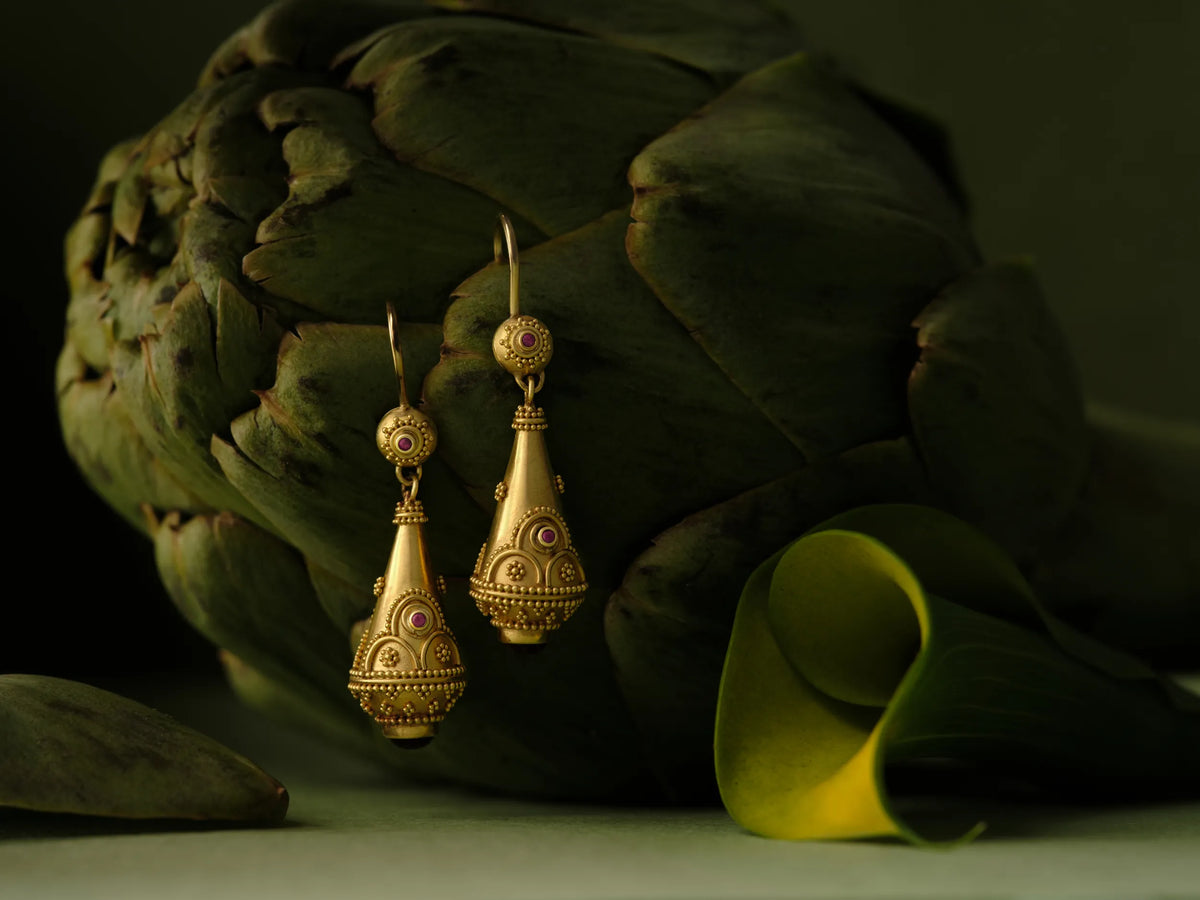 Gold granulated drop earrings set with ruby cabochons, hanging against an artichoke