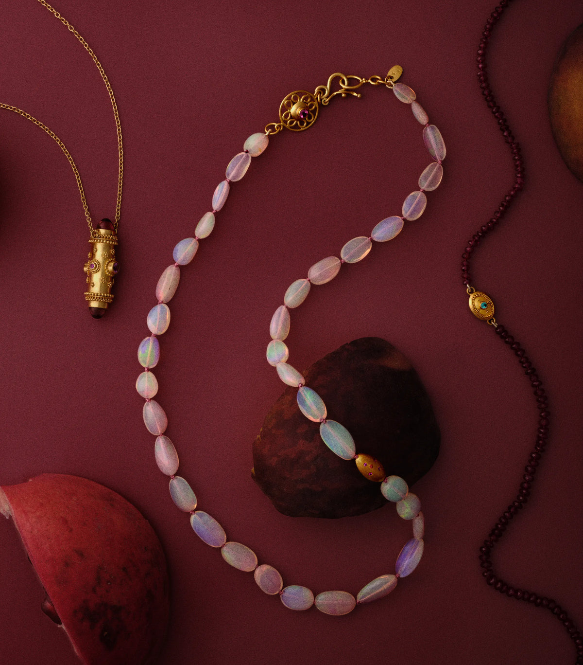 A gold-clasped opal bead necklace with a granulated gold and ruby bead accent, styled beside a garnet bracelet and a garnet cabochon pendant on a deep maroon background