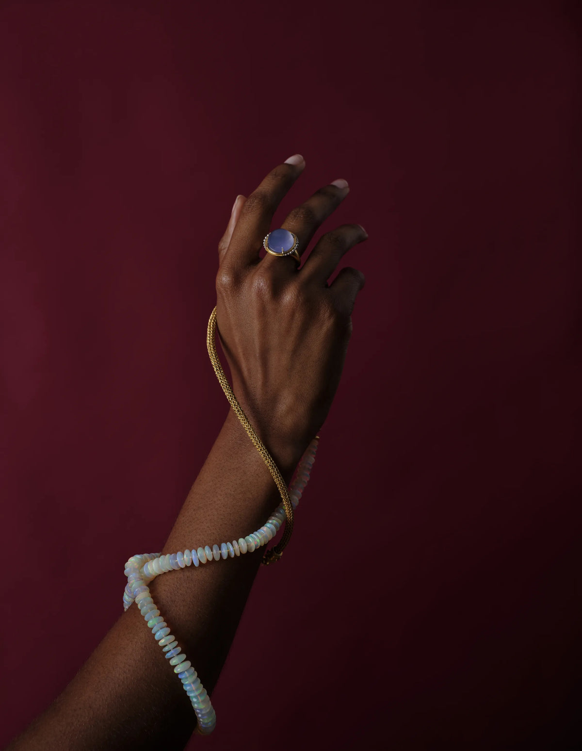 A hand adorned with a gold and blue chalcedony ring, a gold snake chain necklace, and a beaded opal bracelet against a deep maroon background