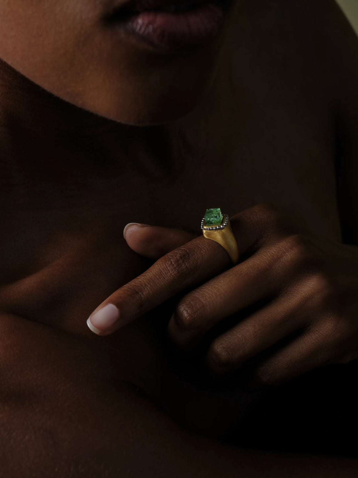 A close-up of a woman's clasped hands wearing a 22k gold ring set with a raw emerald crystal and a beaded border, in low light
