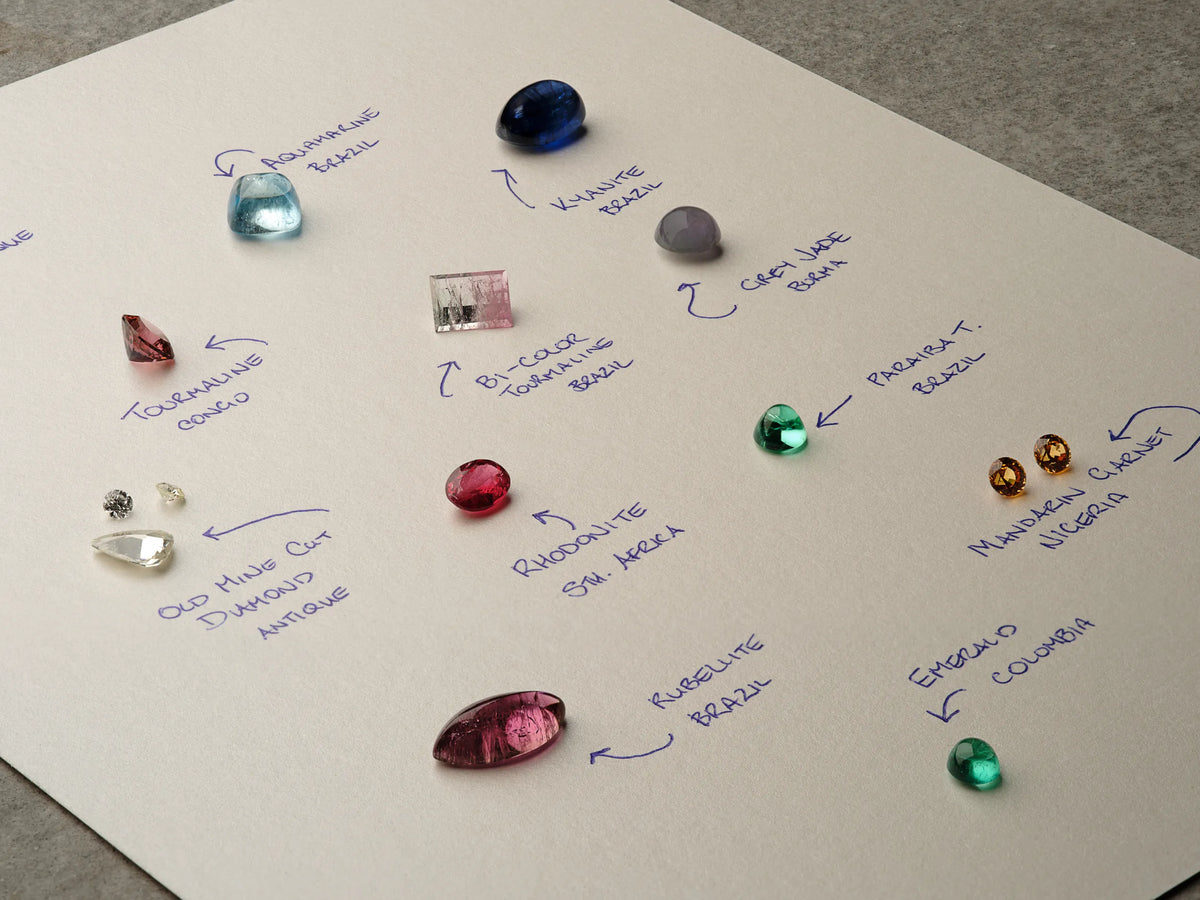 Loose colored gemstones labeled by type and origin, including Paraiba tourmaline from Brazil, Colombian emerald, Nigerian mandarin garnet, and an antique old mine cut diamond